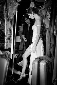 A black and white photograph of a dancer backstage surrounded by props, equipment and two people working